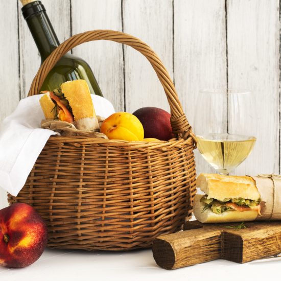 Picnic basket with wine, salmon sandwiches and fruits over white Picnic basket with wine, salmon sandwiches and fruits over white wooden table. Selective focus